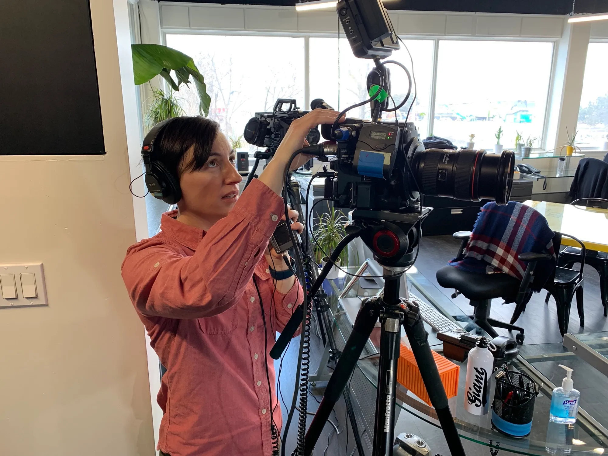 Kate in headphones rigging a cinema camera on a tripod in a bright, plant-filled office, adjusting the audio receiver mounted above the lens