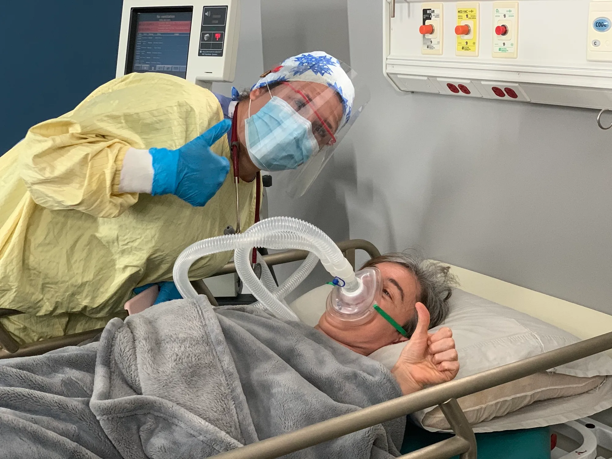 On set in a mock hospital room: a clinician in full PPE beside Kate — playing the patient — who gives a thumbs-up from the bed.
