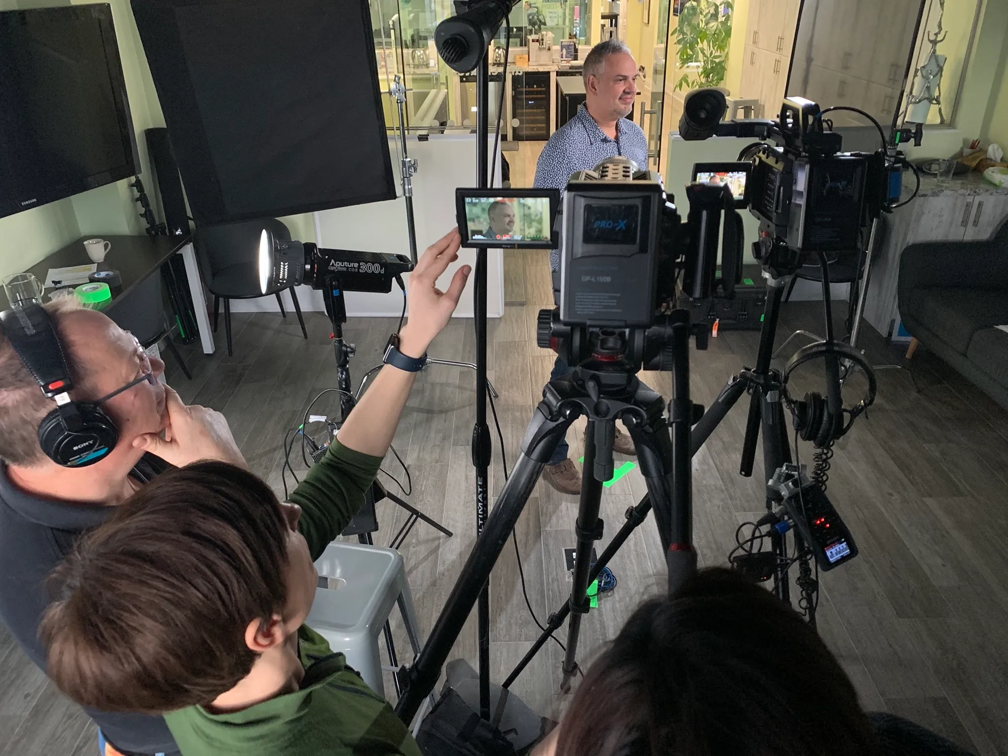 A film crew shooting an interview subject in a bright, plant-filled room, ringed by tripod-mounted cameras, softbox lights and a handheld monitor