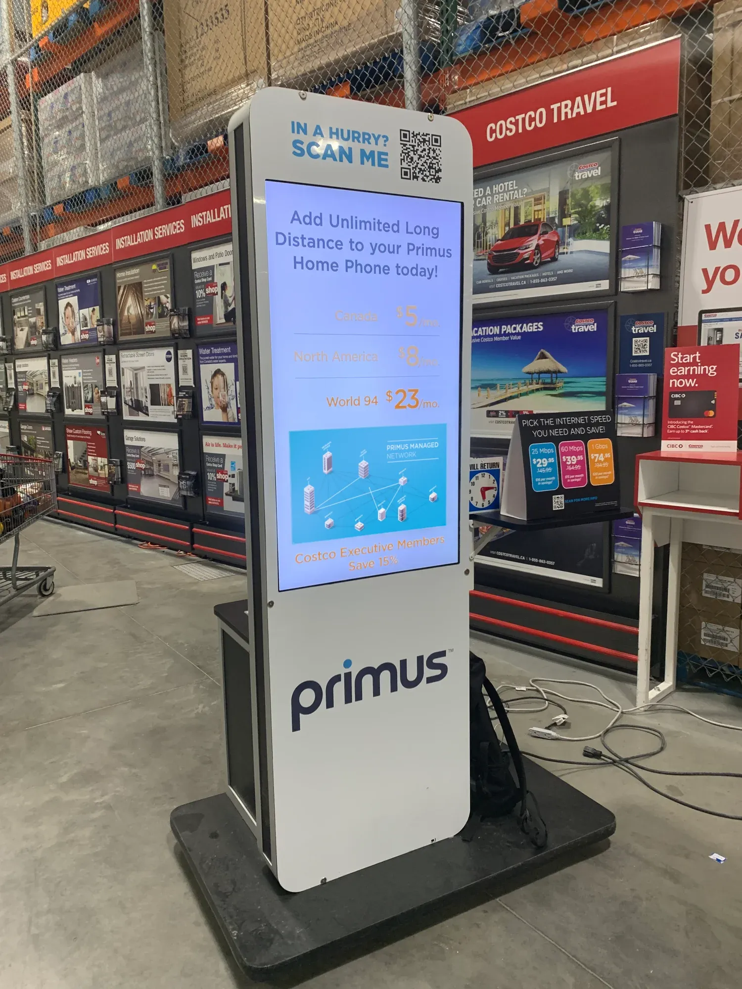 A Primus digital kiosk in a Costco aisle playing the film: the Primus Managed Network frame on screen under long distance pricing, beside the Costco Travel wall.