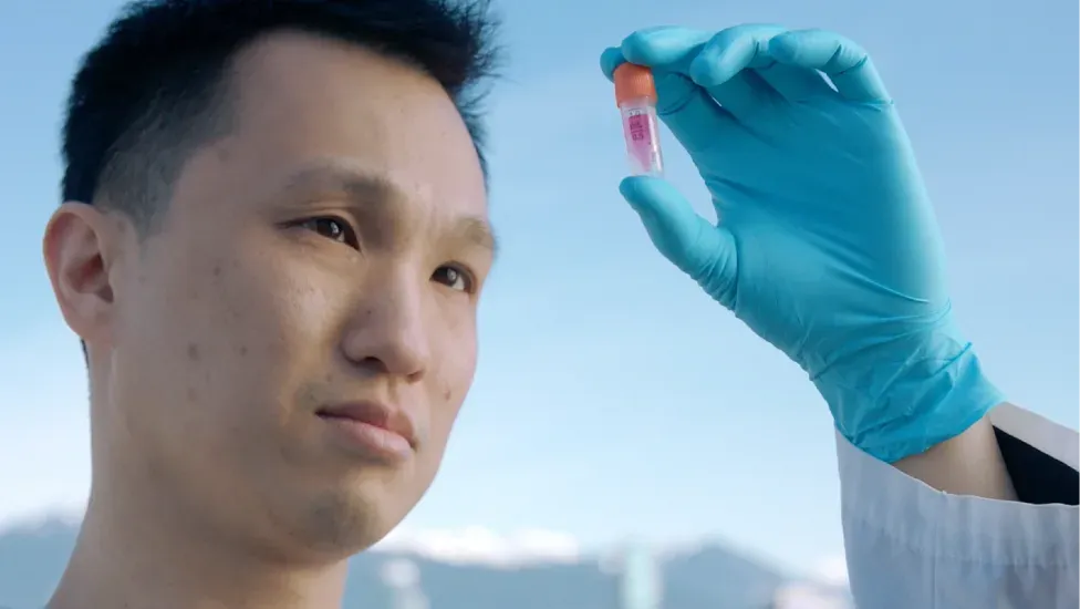 A BC Cancer researcher holds a single-cell sample vial up to the light on a rooftop, the Vancouver skyline and snow-capped mountains behind him.